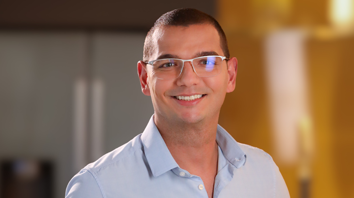 Homem sorrindo, usando óculos de armação transparente e camisa social azul-clara, em retrato com fundo desfocado.