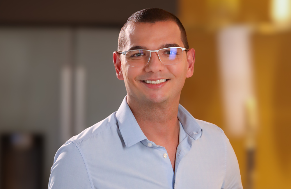 Homem sorrindo, usando óculos de armação transparente e camisa social azul-clara, em retrato com fundo desfocado.