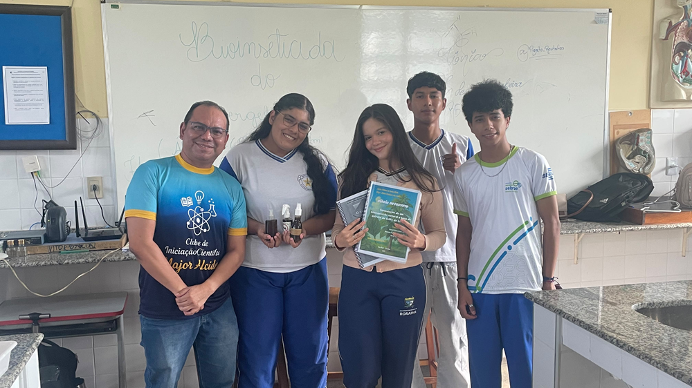 Grupo de estudantes e professor posa junto em sala de aula com quadro branco de fundo