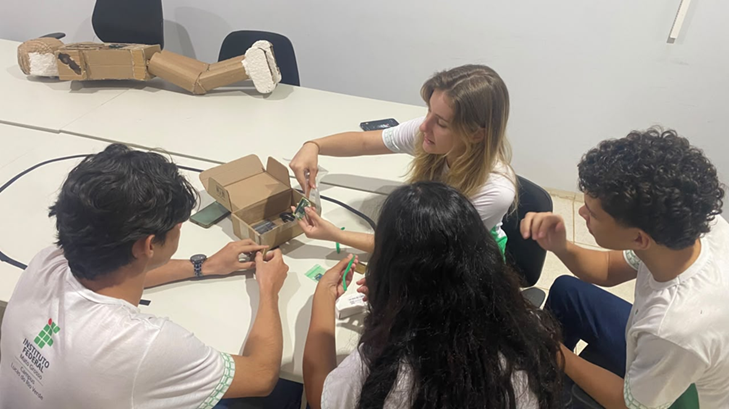 Grupo de dois alunos e duas alunas trabalham juntos em volta de uma mesa manuseando protótipo de visão computacional