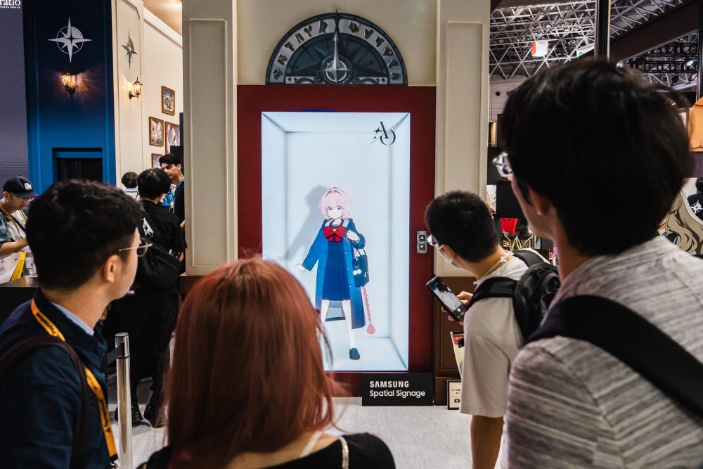 Visitantes reunidos diante de um Samsung Spatial Signage instalado no estande de Astrae Oratio, que exibe uma personagem em uma composição que simula o interior de um elevador.