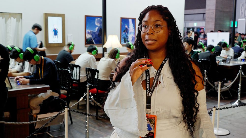 Criadora de conteúdo com crachá da Tokyo Game Show 2026 fala diante da câmera enquanto participantes jogam ao fundo em uma área do estande de Astrae Oratio.
