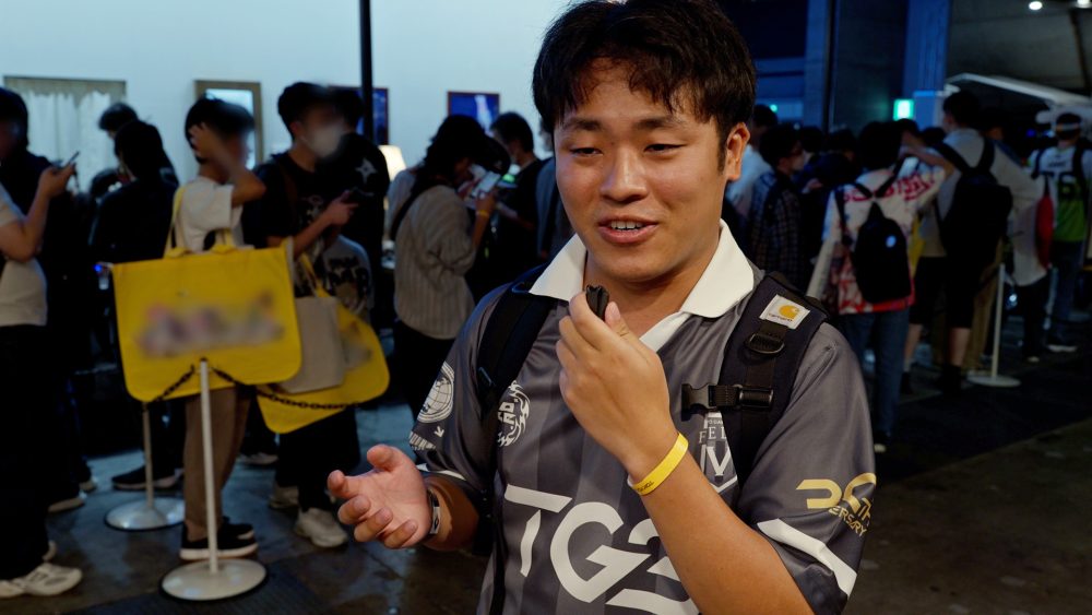 Visitante da Tokyo Game Show 2026 fala diante da câmera enquanto segura um pequeno microfone, com o público e a área do estande de Astrae Oratio ao fundo.