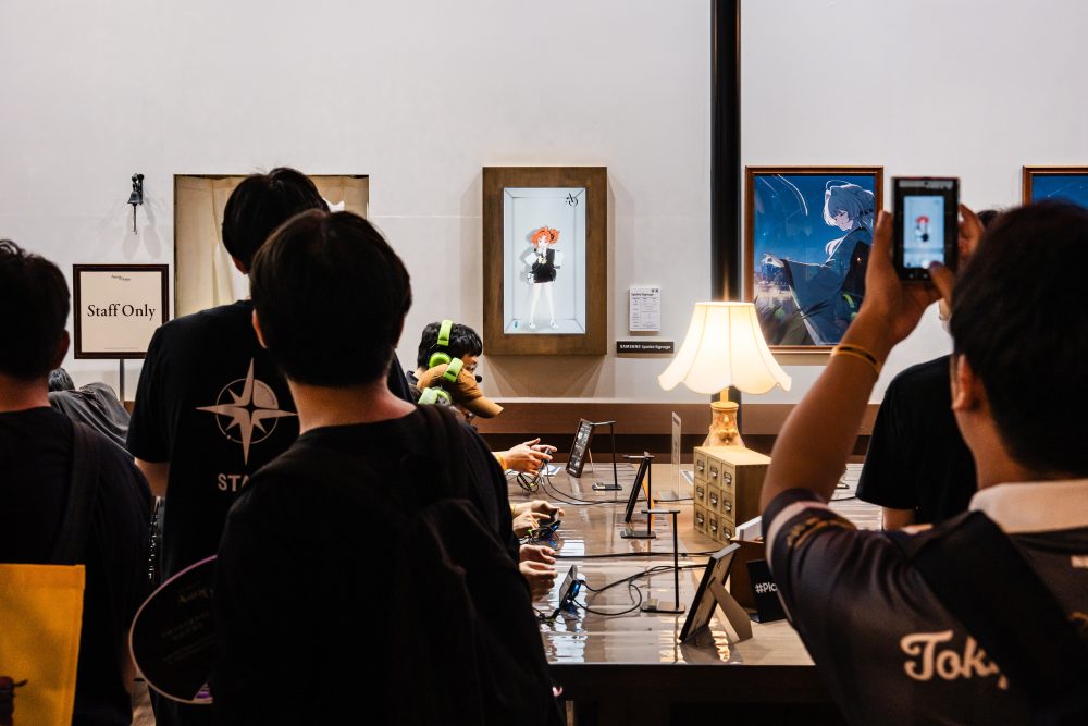 Visitantes observam e fotografam personagens exibidos em Samsung Spatial Signage instalados em uma parede do estande de Astrae Oratio, enquanto outras pessoas jogam em uma área de atendimento ao fundo.