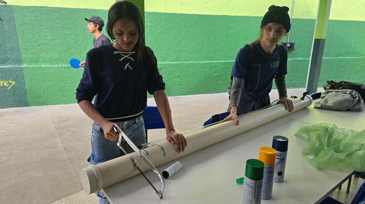 Um aluno e uma aluna na área externa de escola cortando um cano para projeto científico