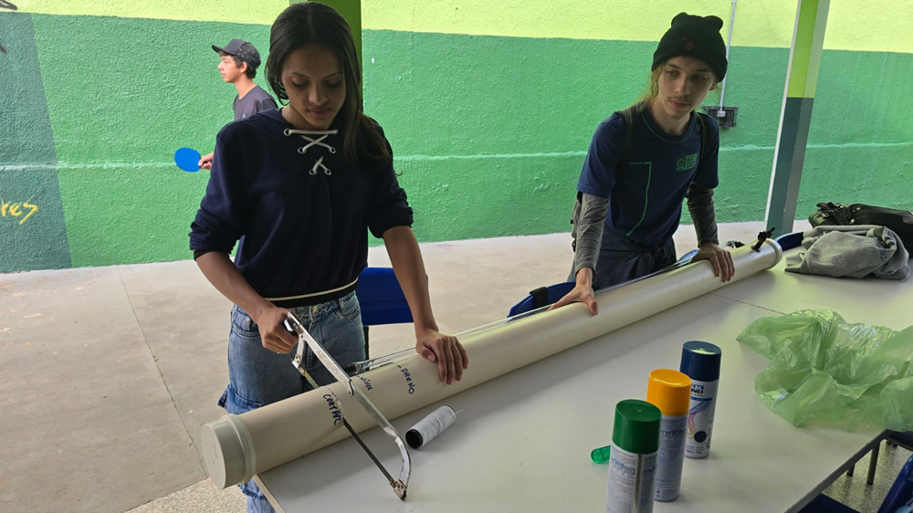 Um aluno e uma aluna na área externa de escola cortando um cano para projeto científico