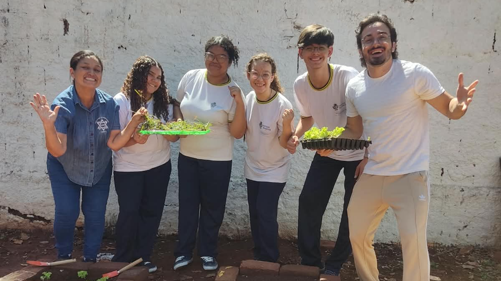 Grupo de quatro alunos e dois professores posam juntos em horta escolar com plantas nas mãos
