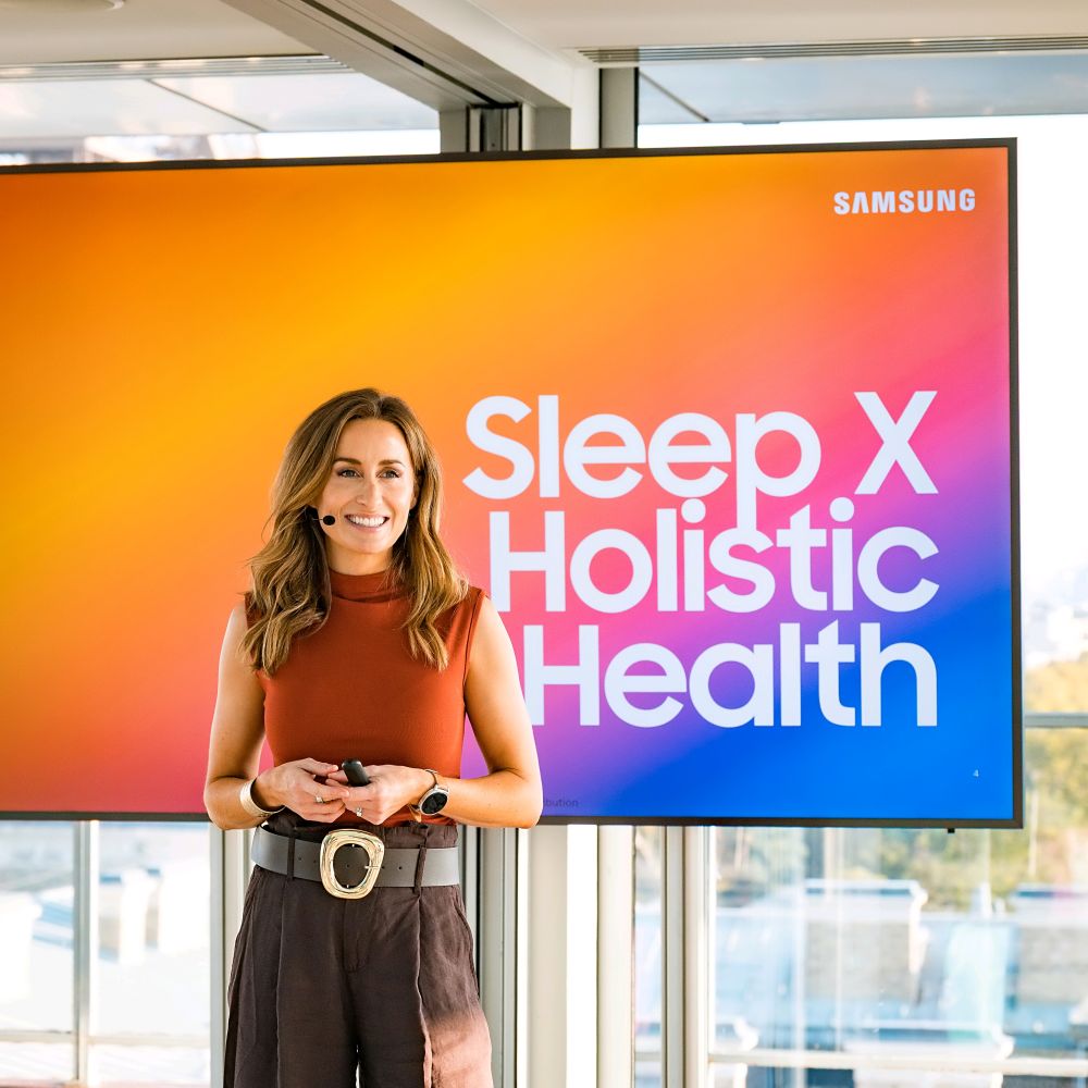 Frau spricht mit Mikrofon vor einer Präsentationsfolie mit der Aufschrift „Sleep X Holistic Health“ von Samsung.