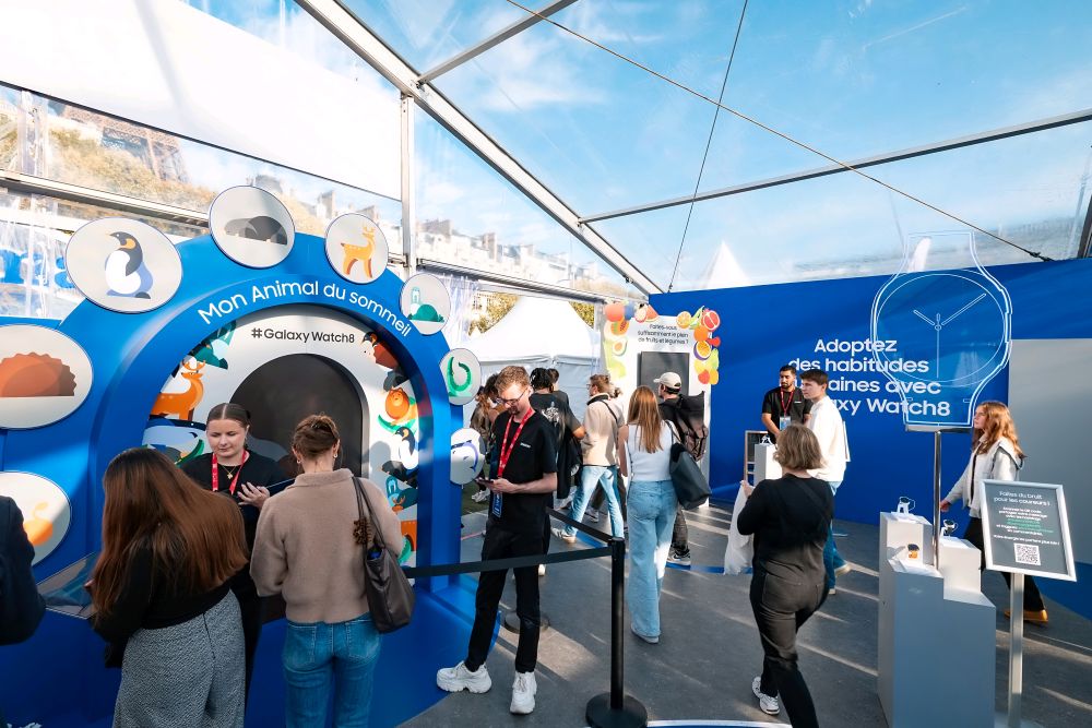 Besucherinnen und Besucher erkunden einen farbenfrohen Samsung-Stand in einem Zelt mit transparentem Dach. Wände und Displays zeigen Illustrationen und Texte rund um die Galaxy Watch und Schlafgewohnheiten, während Menschen interaktiv verschiedene Stationen ausprobieren.