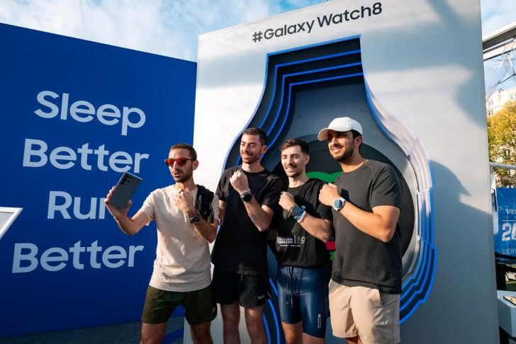 Vier Männer posieren vor einer Wand mit der Aufschrift „Sleep Better, Run Better“ und dem Hashtag „#GalaxyWatch8“. Sie tragen Sportkleidung und Smartwatches und machen gemeinsam ein Selfie vor einem großen Display in Form einer Uhr.