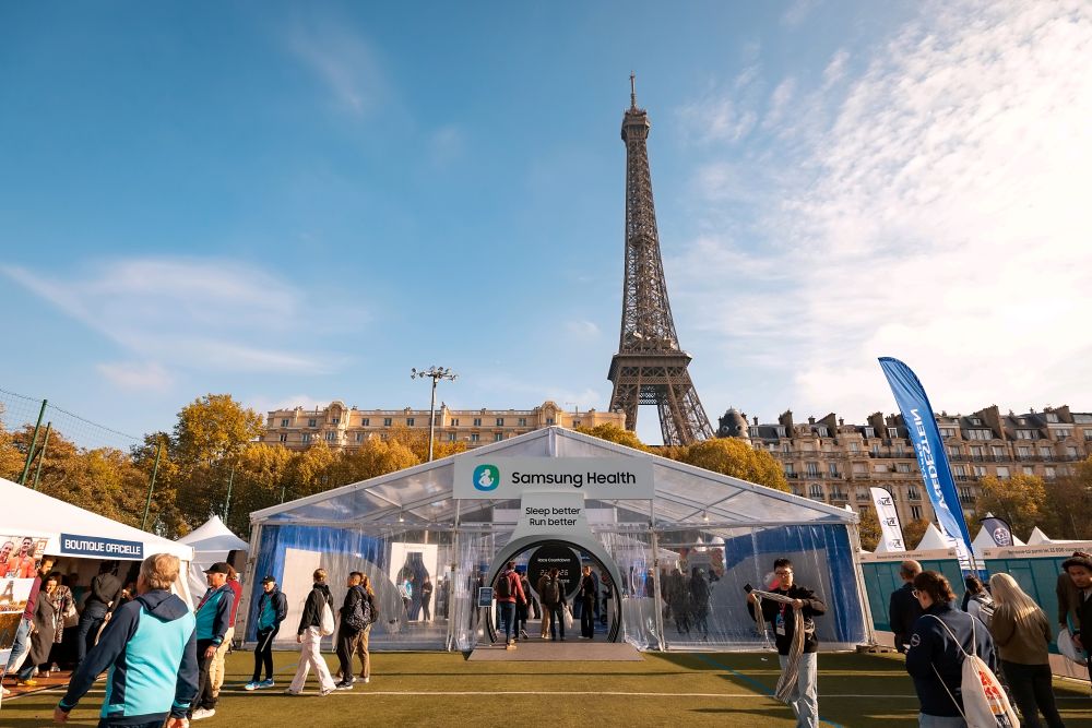 Menschen besuchen ein großes Zelt mit der Aufschrift „Samsung Health“ vor dem Eiffelturm in Paris. Das sonnige Wetter und die umliegenden Gebäude schaffen eine lebendige, einladende Atmosphäre.