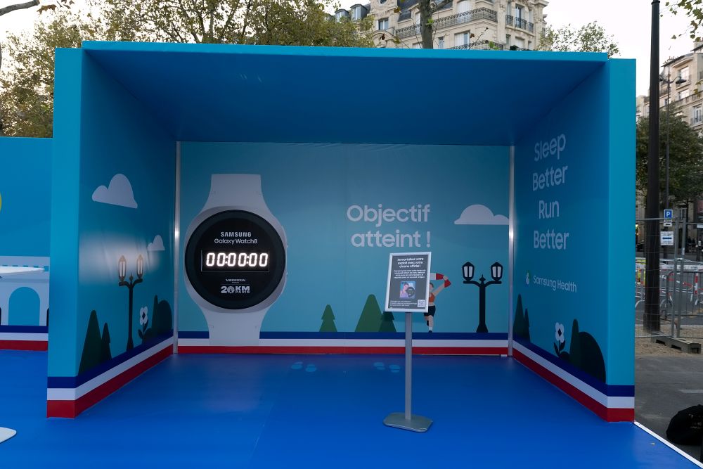 Eine Ausstellungsfläche mit blauem Hintergrund zeigt eine große Darstellung einer Samsung Galaxy Watch mit der Aufschrift „Objectif atteint!“ und „Sleep Better Run Better“. Die Gestaltung erinnert an eine Lauf- oder Fitnesskampagne im Freien.