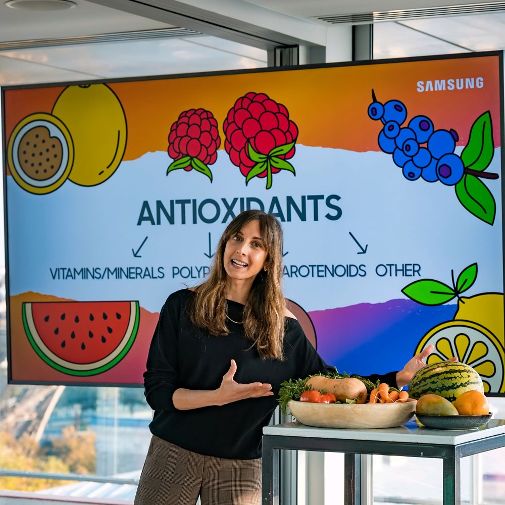 Vortragende spricht über gesunde Ernährung vor einer farbenfrohen Präsentationsfolie mit Illustrationen von Früchten.