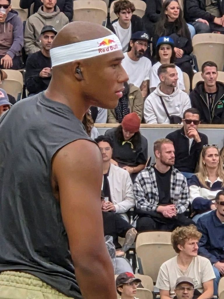 Ein Sportler mit einem weißen Red-Bull-Stirnband und kabellosen Ohrhörern steht fokussiert vor einer Zuschauermenge. Im Hintergrund sitzen zahlreiche Menschen auf Tribünen und beobachten das Geschehen aufmerksam.