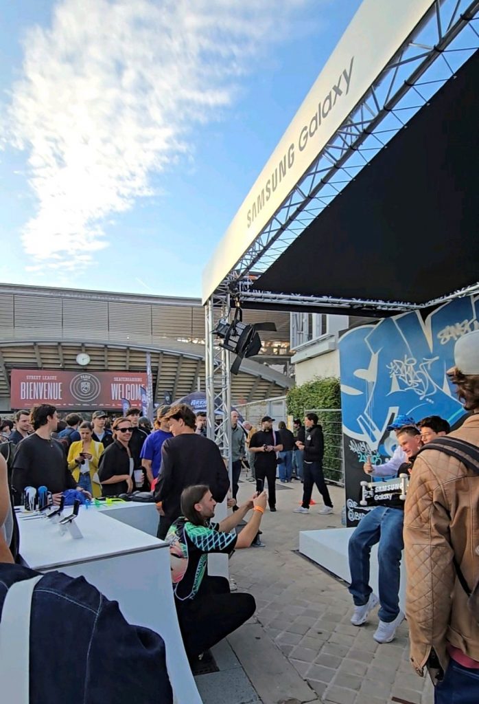 Eine Menschenmenge versammelt sich vor einem überdachten Samsung-Galaxy-Bereich im Freien. Einige Besucher sitzen und machen Fotos, während andere vor einer blauen Wand mit Graffiti stehen. Im Hintergrund sind Banner mit der Aufschrift „Bienvenue“ und „Welcome“ zu sehen.