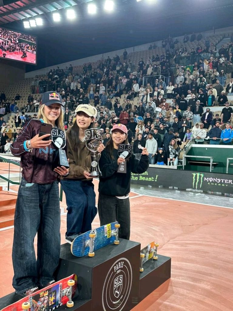Drei Skateboarderinnen posieren mit SLS-Pokalen und -Trophäen vor einer großen Zuschauermenge in einem beleuchteten Stadion. Sie lächeln in die Kamera, während im Hintergrund das Publikum jubelt.