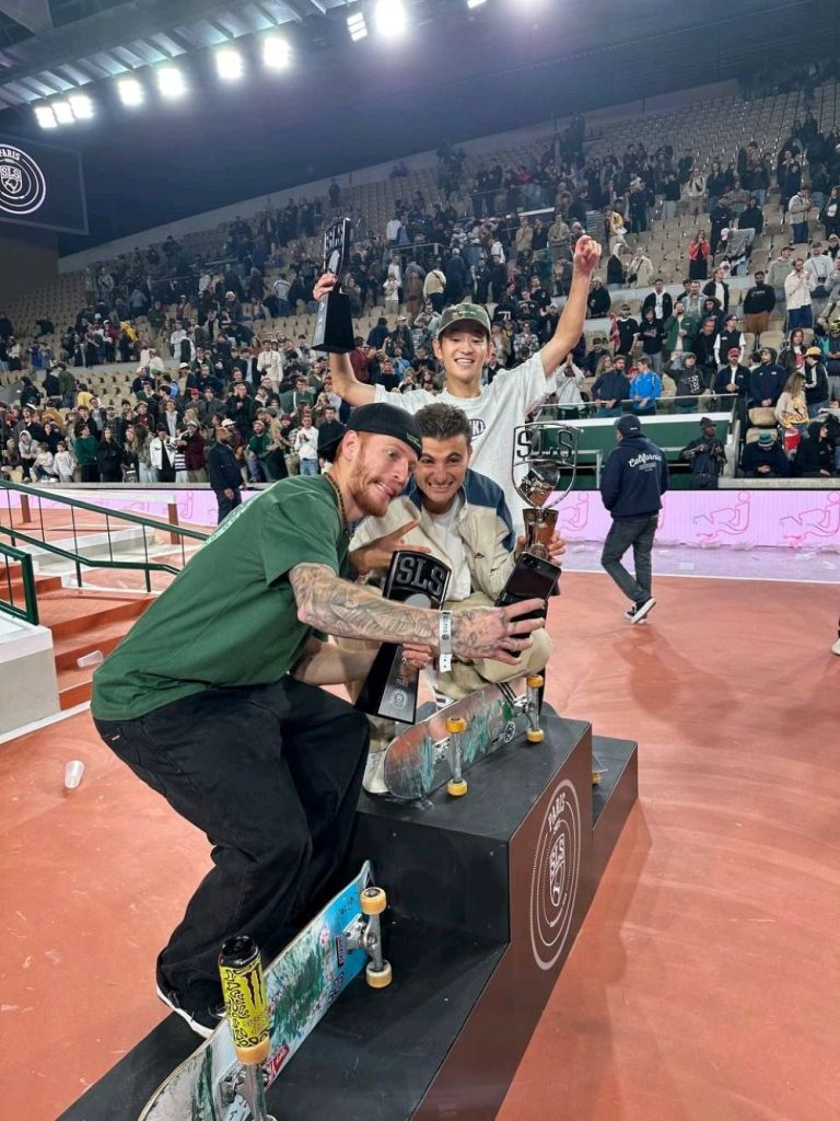 Drei Skateboarder posieren mit SLS-Pokalen auf einem Podest in einer Skatehalle. Sie lächeln und halten ihre Trophäen, während im Hintergrund Zuschauer auf den Tribünen jubeln. Zwei Skateboards stehen vor ihnen auf dem Podest.