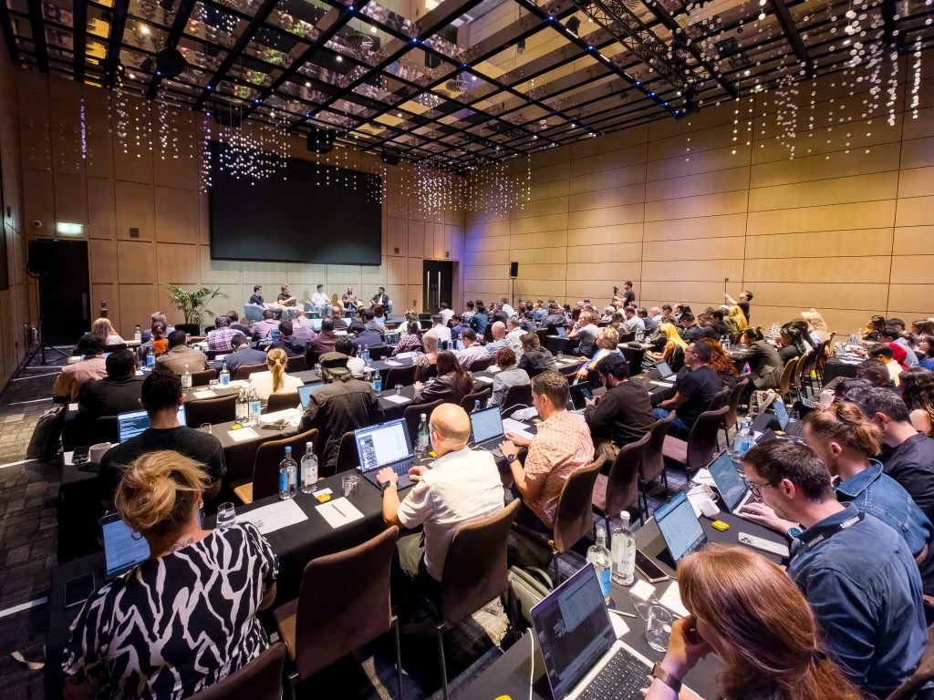 Weitwinkelfoto eines gut besuchten Konferenzsaals: Zahlreiche Teilnehmerinnen und Teilnehmer sitzen an runden Tischen mit Laptops und Wasserflaschen und verfolgen eine Podiumsdiskussion auf der beleuchteten Bühne im Hintergrund. Das Bild zeigt das Health Forum im Rahmen von Samsung Galaxy Unpacked 2026 in London.