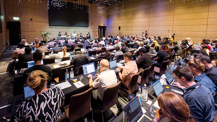 Weitwinkelfoto eines gut besuchten Konferenzsaals: Zahlreiche Teilnehmerinnen und Teilnehmer sitzen an runden Tischen mit Laptops und Wasserflaschen und verfolgen eine Podiumsdiskussion auf der beleuchteten Bühne im Hintergrund. Das Bild zeigt das Health Forum im Rahmen von Samsung Galaxy Unpacked 2026 in London.