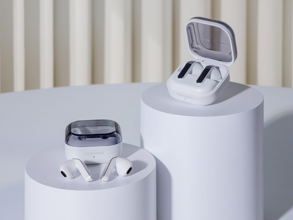 Zwei Paar weiße Samsung Galaxy Buds4 Pro und Galaxy Buds4 mit geöffneten Ladeetuis stehen auf weißen Podesten vor einem hellen Hintergrund.