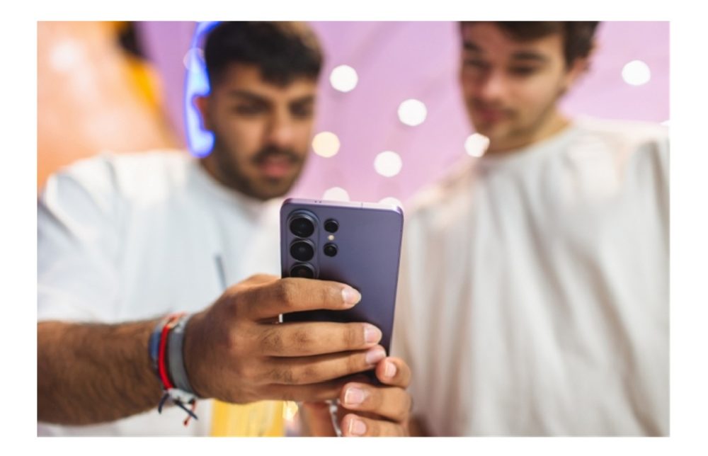 Zwei Männer betrachten gemeinsam den Bildschirm eines Samsung Galaxy S26 Ultra vor einem farbenfrohen, unscharfen Hintergrund. Einer der Männer umfasst das Smartphone mit seinen Händen. Sichtbar ist die Rückseite des Samsung Galaxy S26 Ultra in der Farbe Cobalt Violet.