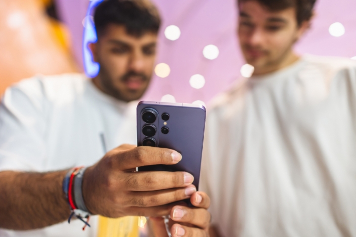 Zwei Männer betrachten gemeinsam den Bildschirm eines Samsung Galaxy S26 Ultra vor einem farbenfrohen, unscharfen Hintergrund. Einer der Männer umfasst das Smartphone mit seinen Händen. Sichtbar ist die Rückseite des Samsung Galaxy S26 Ultra in der Farbe Cobalt Violet.