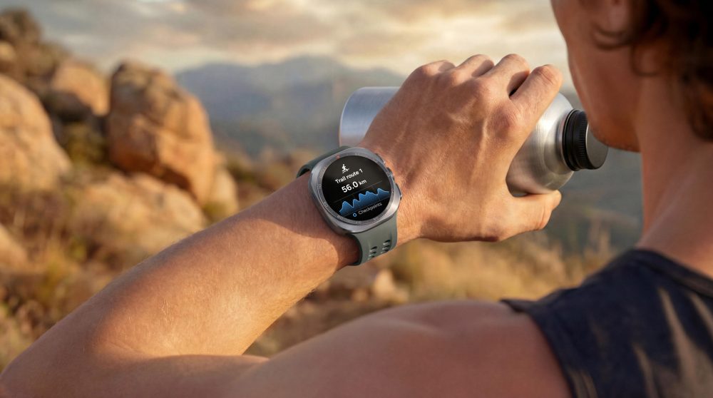 Person von hinten mit Samsung Galaxy Watch Ultra2 am Handgelenk, die aus einer Metallflasche trinkt. Im Hintergrund eine felsige Landschaft. Auf dem Display der Uhr ist die Trail-Route mit der Streckenlänge und einem Höhenprofil zu sehen.