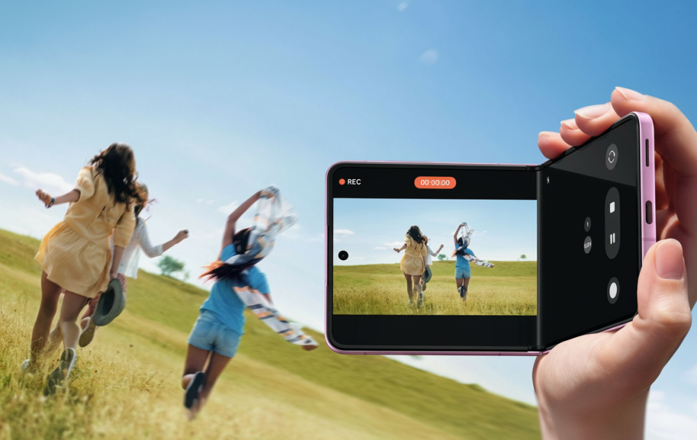 Eine Frauenhand hält ein rechtwinklig aufgeklapptes Samsung Galaxy Z Flip8. Auf dem Display und im Hintergrund sieht man drei Frauen, die über eine Wiese laufen.