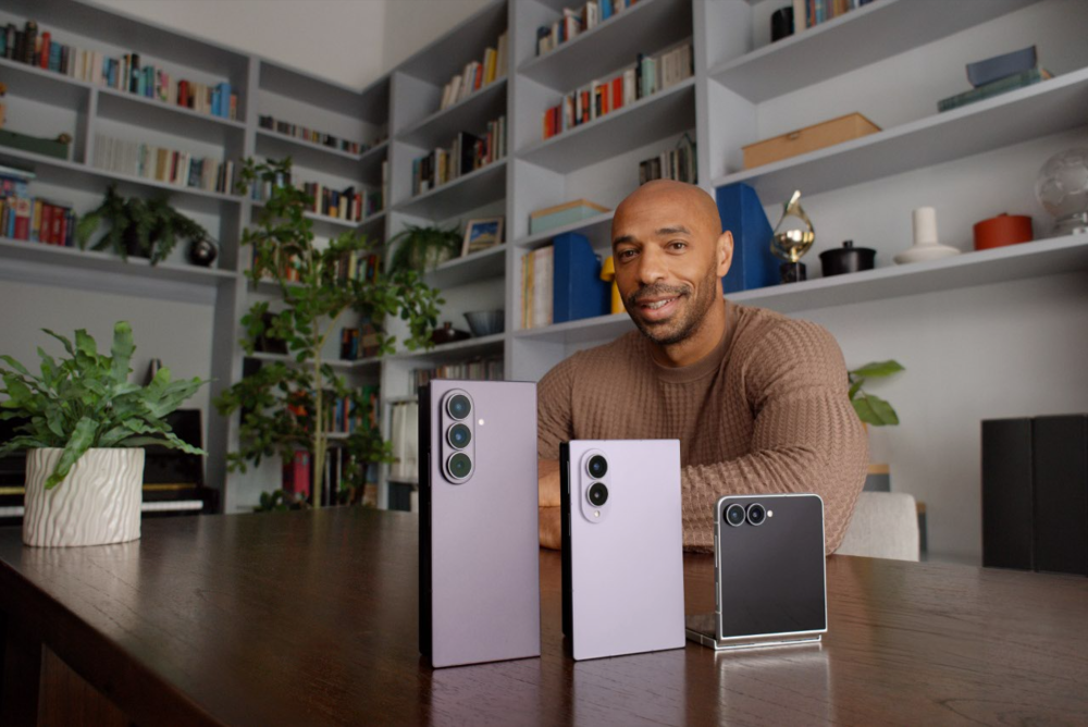 Der ehemalige Fußballstar Thierry Henry sitzt an einem Tisch vor einem Bücherregal. Vor ihm stehen die drei Geräte der Samsung Galaxy Z8-Serie.