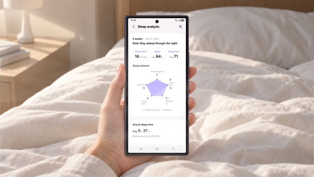 Eine Hand hält ein Samsung Galaxy Smartphone mit der Schlafanalyse in der Samsung Health-App, im Hintergrund ein weiß bezogenes Bett im warmen Morgenlicht.