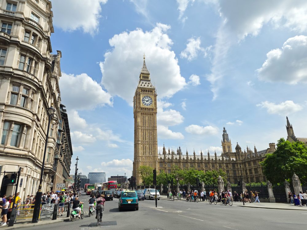 Elizabeth Tower and the Palace of Westminster in London, seen from a busy street with pedestrians, cyclists and vehicles under a blue sky.