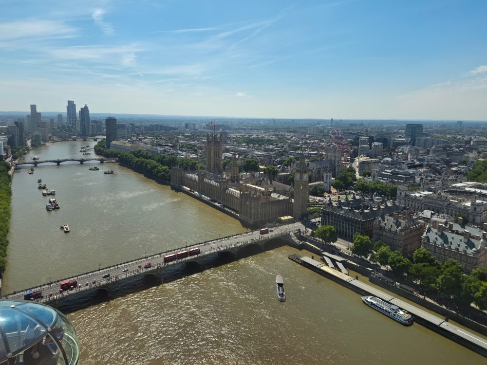 Aerial view of the River Thames in London, with Westminster Bridge, the Palace of Westminster and Elizabeth Tower visible beside the river under a clear blue sky.