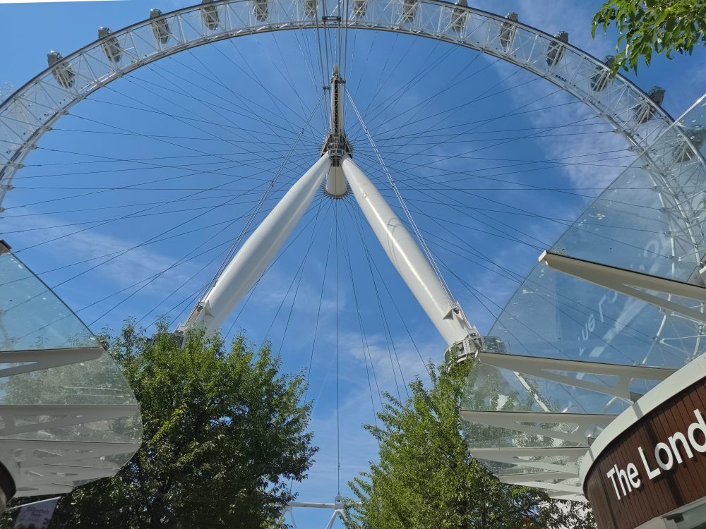 Close-up view of the London Eye from below, showing its white support structure, cables and passenger capsules against a clear blue sky.