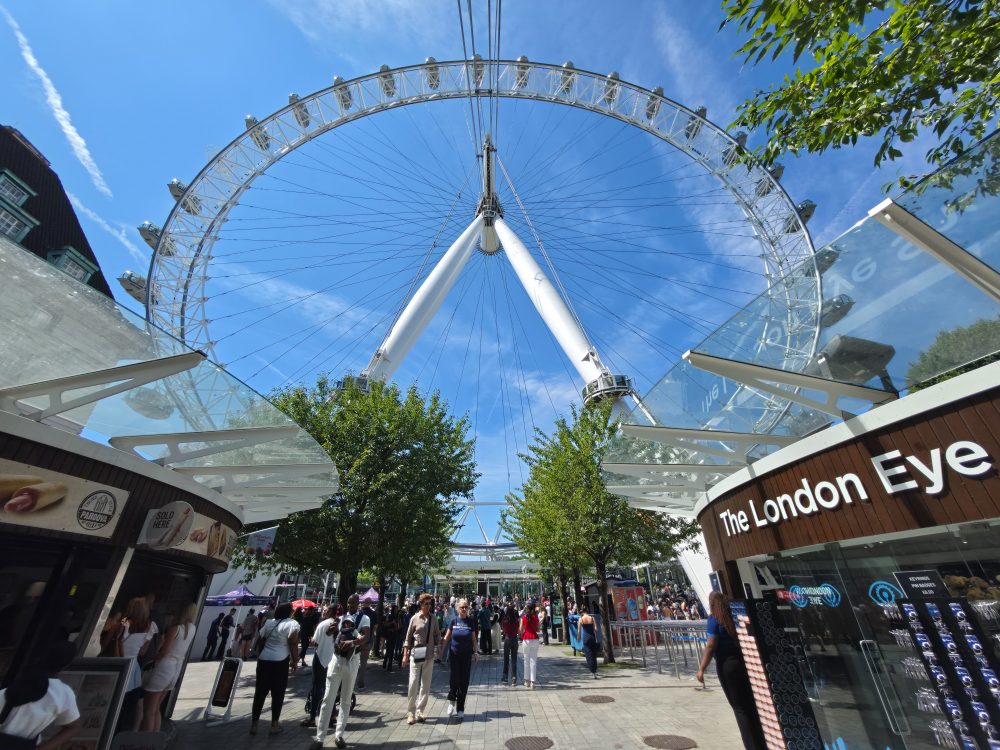 The London Eye observation wheel viewed from below, with visitors walking through the entrance area beneath a clear blue sky.