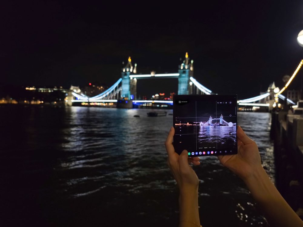 A person photographs Tower Bridge at night with an unfolded Samsung Galaxy device, while the illuminated bridge appears across the River Thames.