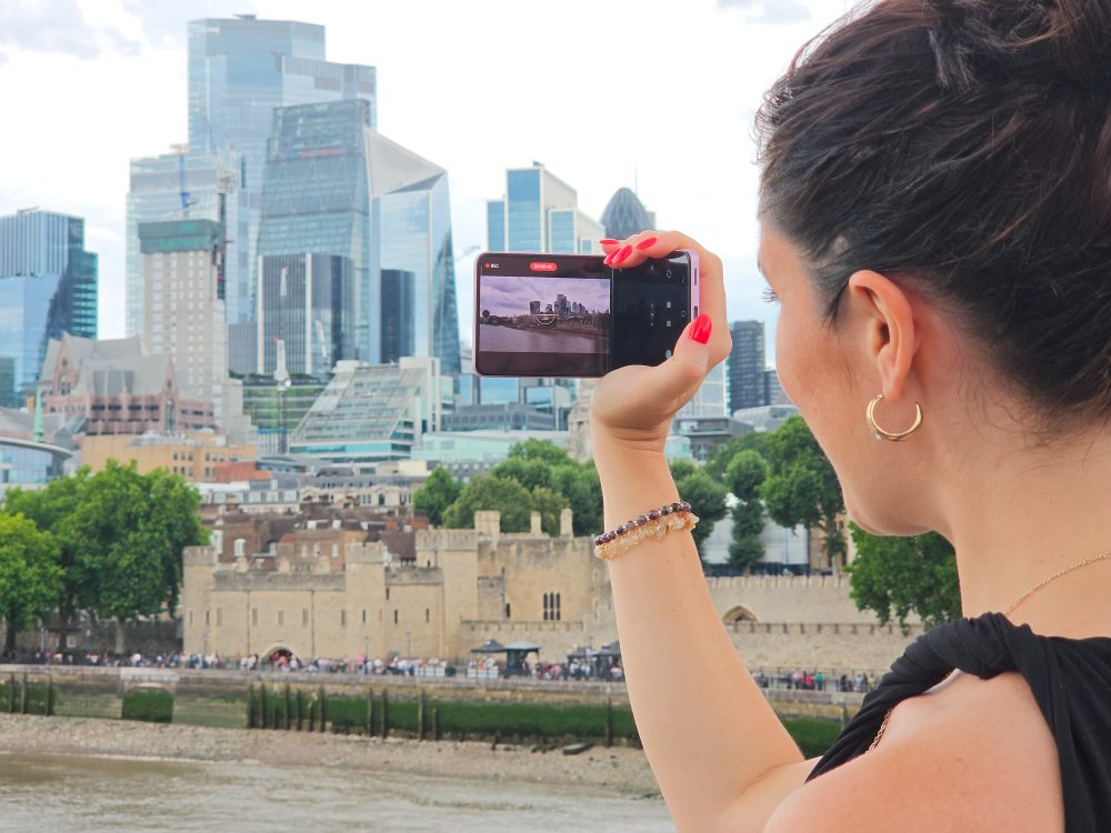 A woman records the London skyline with a Samsung Galaxy foldable smartphone held horizontally, with the Tower of London and City of London in the background.
