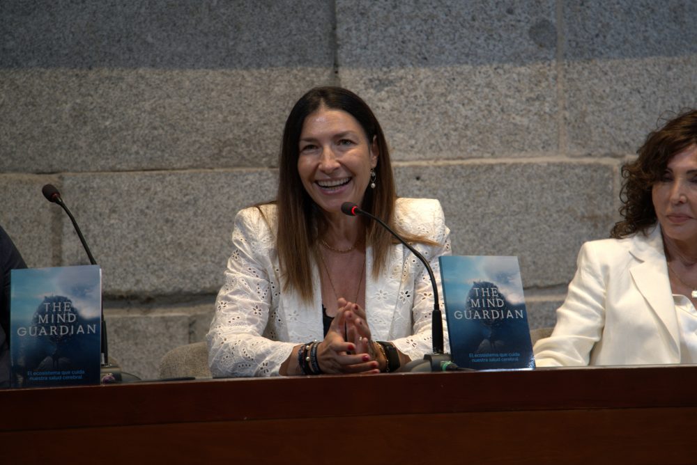 Mujer interviene ante un micrófono durante la presentación de The Mind Guardian: El ecosistema que cuida nuestra salud cerebral, con ejemplares del libro sobre la mesa.