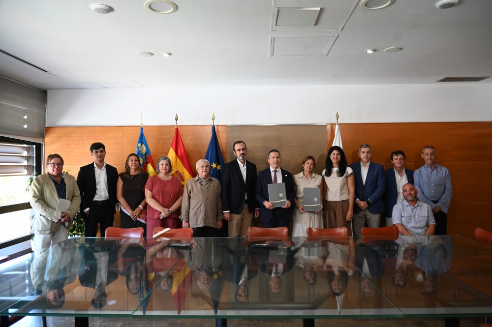 Grupo de representantes posa tras la firma de un acuerdo en la Universitat Politècnica de València, con las banderas de la Comunidad Valenciana, España, la Unión Europea y la UPV al fondo.