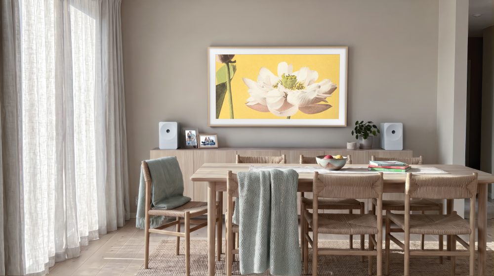Comedor luminoso de estilo contemporáneo, con mesa y sillas de madera, aparador, altavoces blancos y un televisor de gran formato que muestra una flor blanca sobre fondo amarillo.