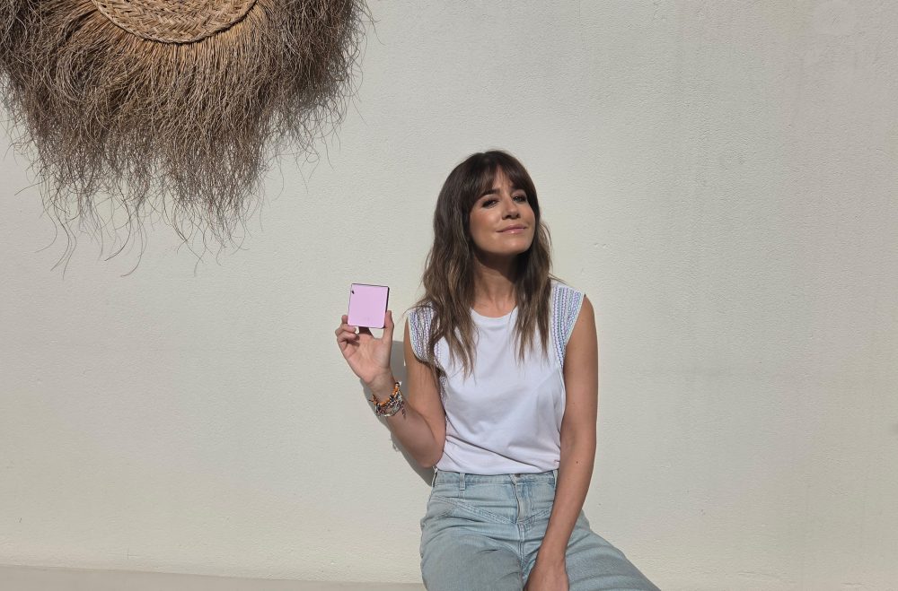 Mujer sosteniendo un Samsung Galaxy Z Flip de color rosa frente a una pared blanca, en un entorno exterior.