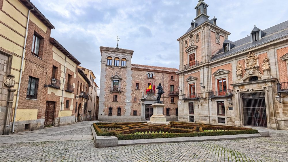 Vista de la Plaza de la Villa, en el Madrid de los Austrias, con la estatua de Álvaro de Bazán, jardines geométricos y edificios históricos de ladrillo y piedra.