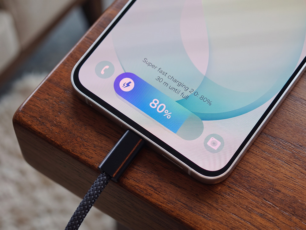 Galaxy S26 FE di atas meja kayu sedang diisi daya dengan kabel. Layar menunjukkan Super Fast Charging dengan baterai 80%.