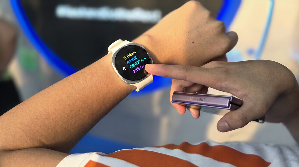 Pelari menunjukkan Galaxy Watch9 berwarna putih yang menampilkan data lari 5,04 km, waktu 41:02, dan pace 8'07" per km, sambil memegang Galaxy Z Flip8 di Galaxy Runners Station